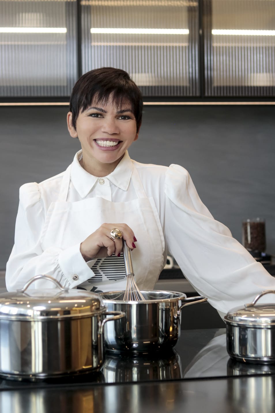 Chef Maria José sorrindo na cozinha ao lado de panelas de aço inoxidável